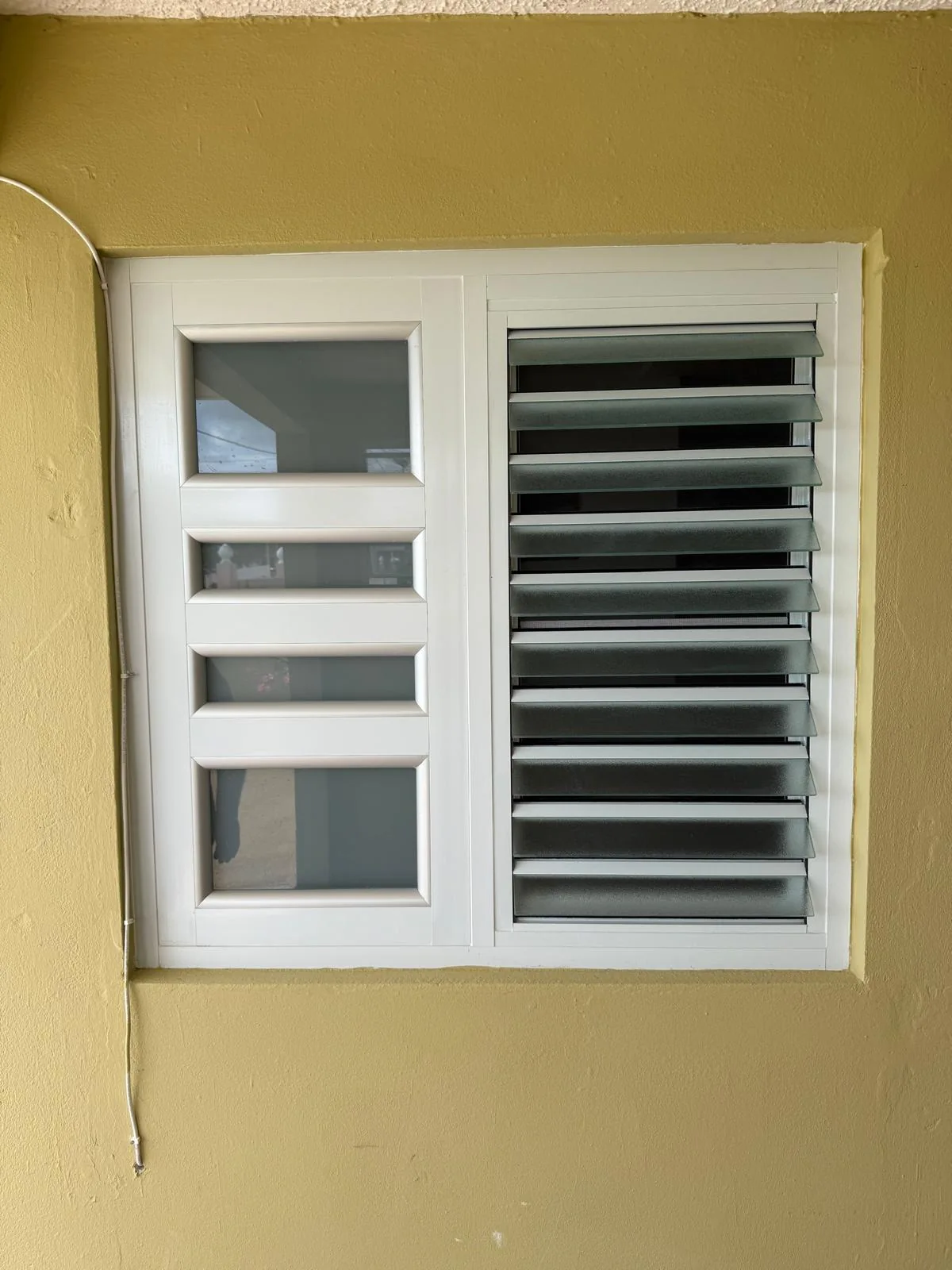 Ventana blanca de aluminio con panel de cristal y lamas, instalada en pared amarilla.